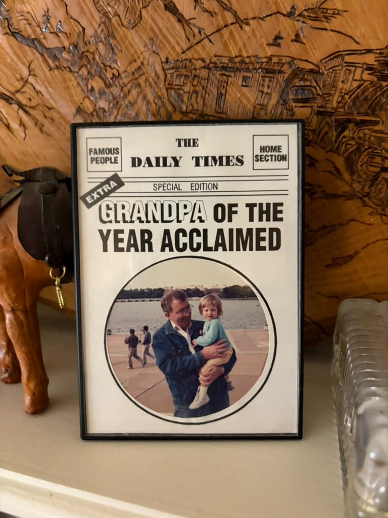A framed Grandpa of the Year keepsake on a mantel with carved wood and surrounding objects